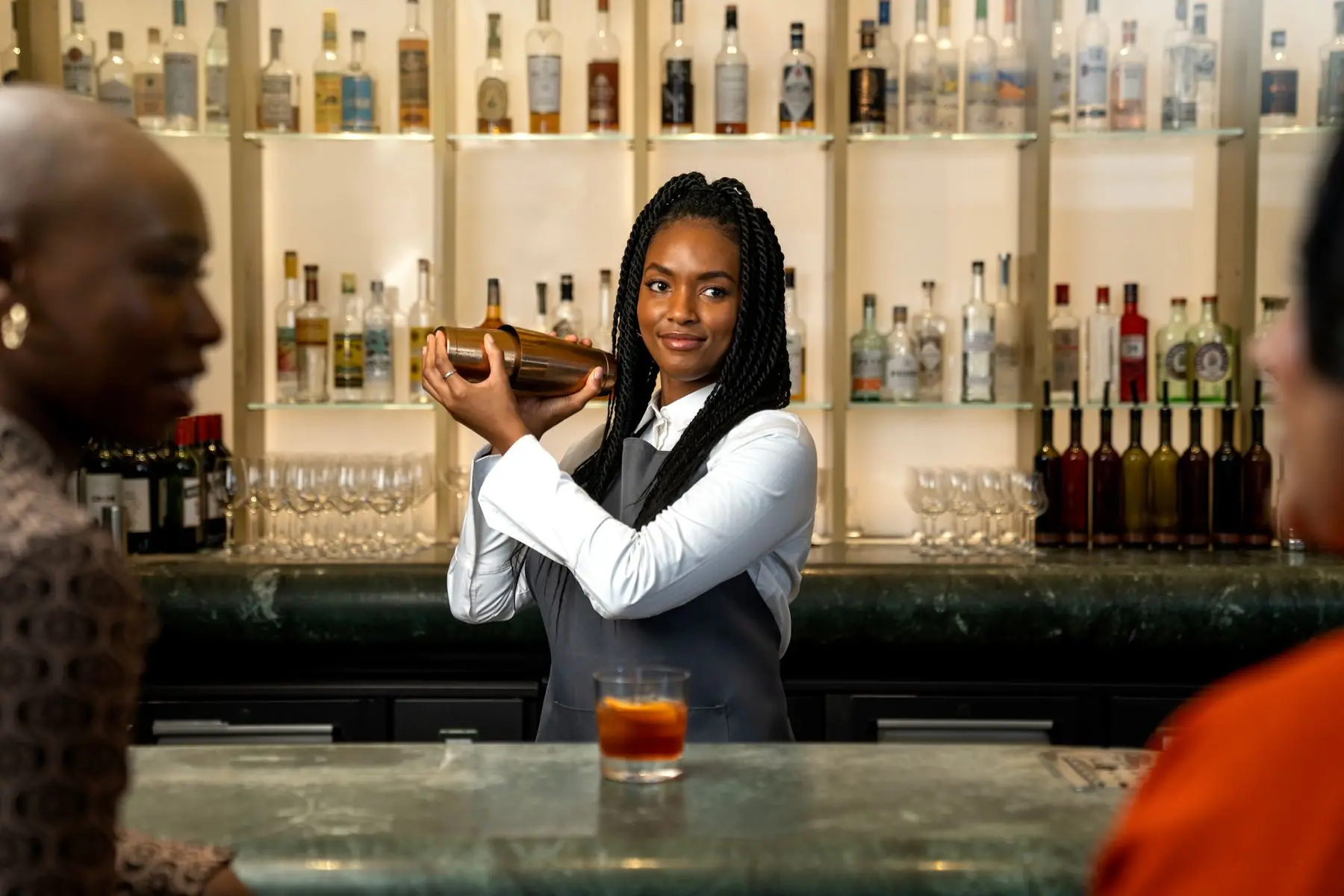 Barmaid agitant un shaker au comptoir d'un bar à cocktails, devant des étagères de bouteilles.