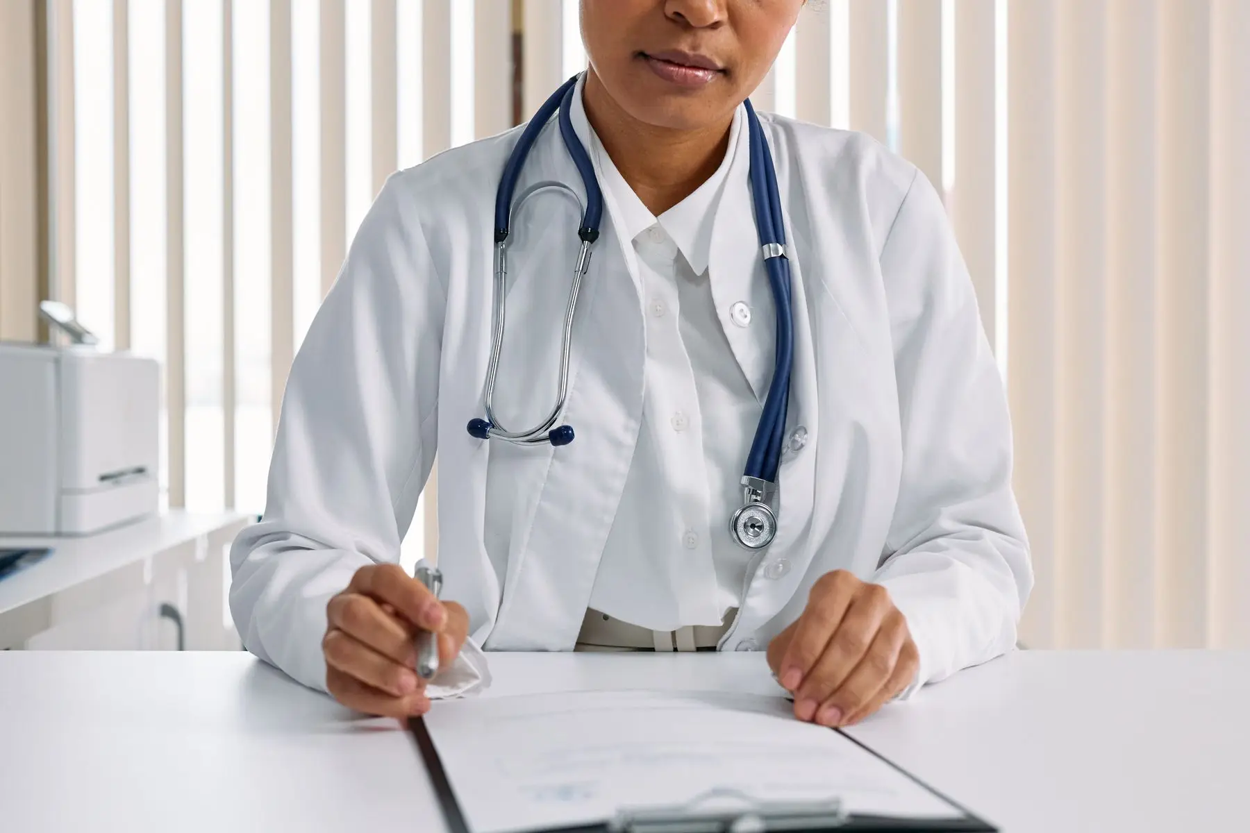 Une médecin en blouse blanche écrit à son bureau, un stéthoscope autour du cou.