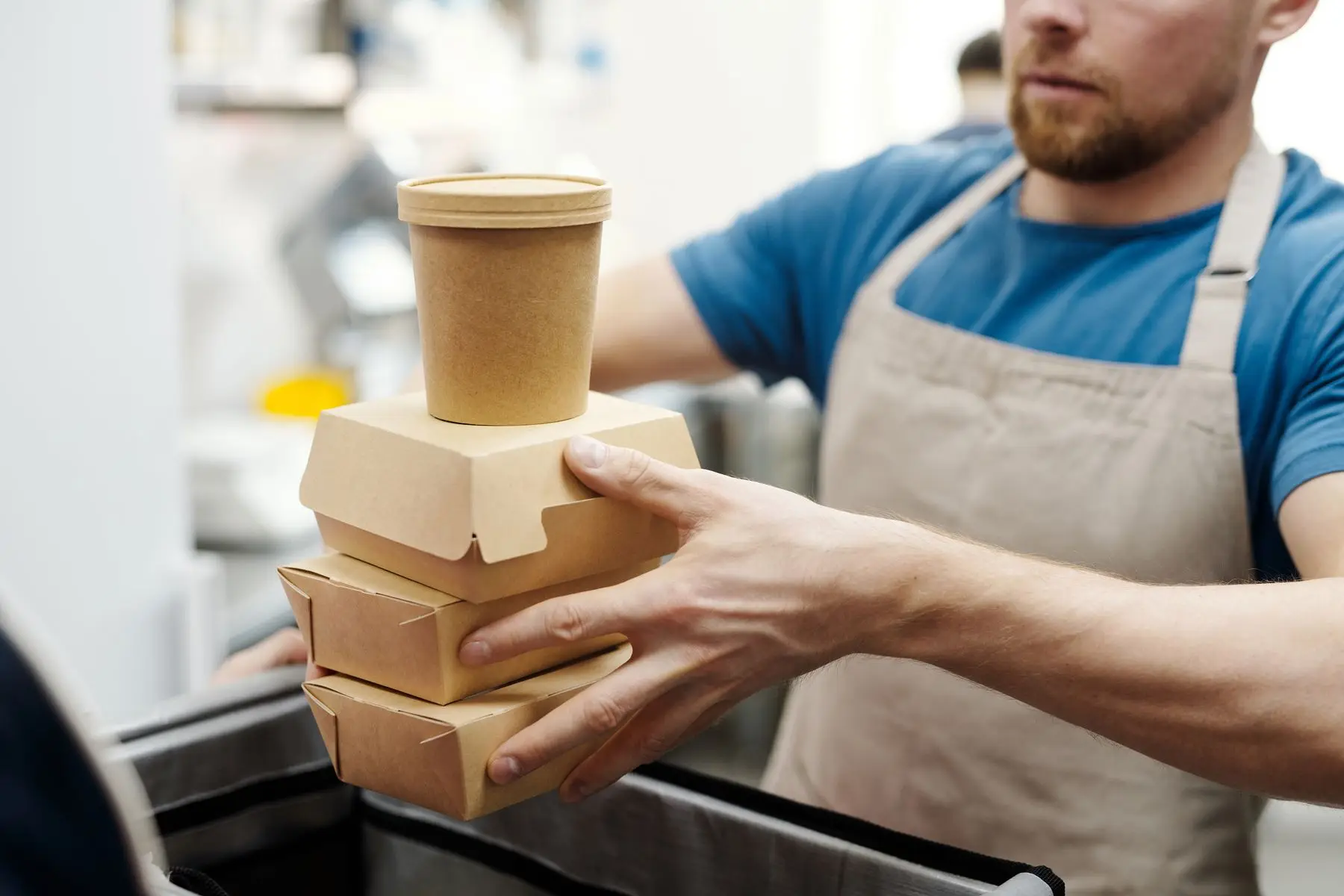 Un cuisinier en tablier tient des boîtes à emporter en carton au-dessus d'un sac isotherme.