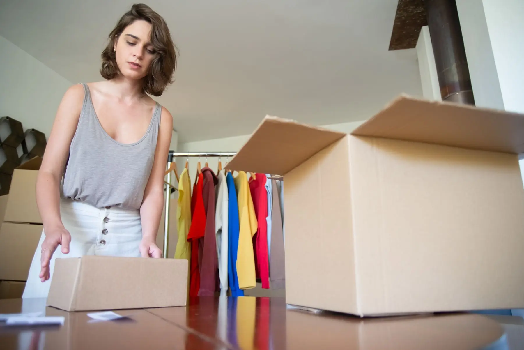 Une jeune femme ouvre des cartons devant un portant de vêtements colorés.