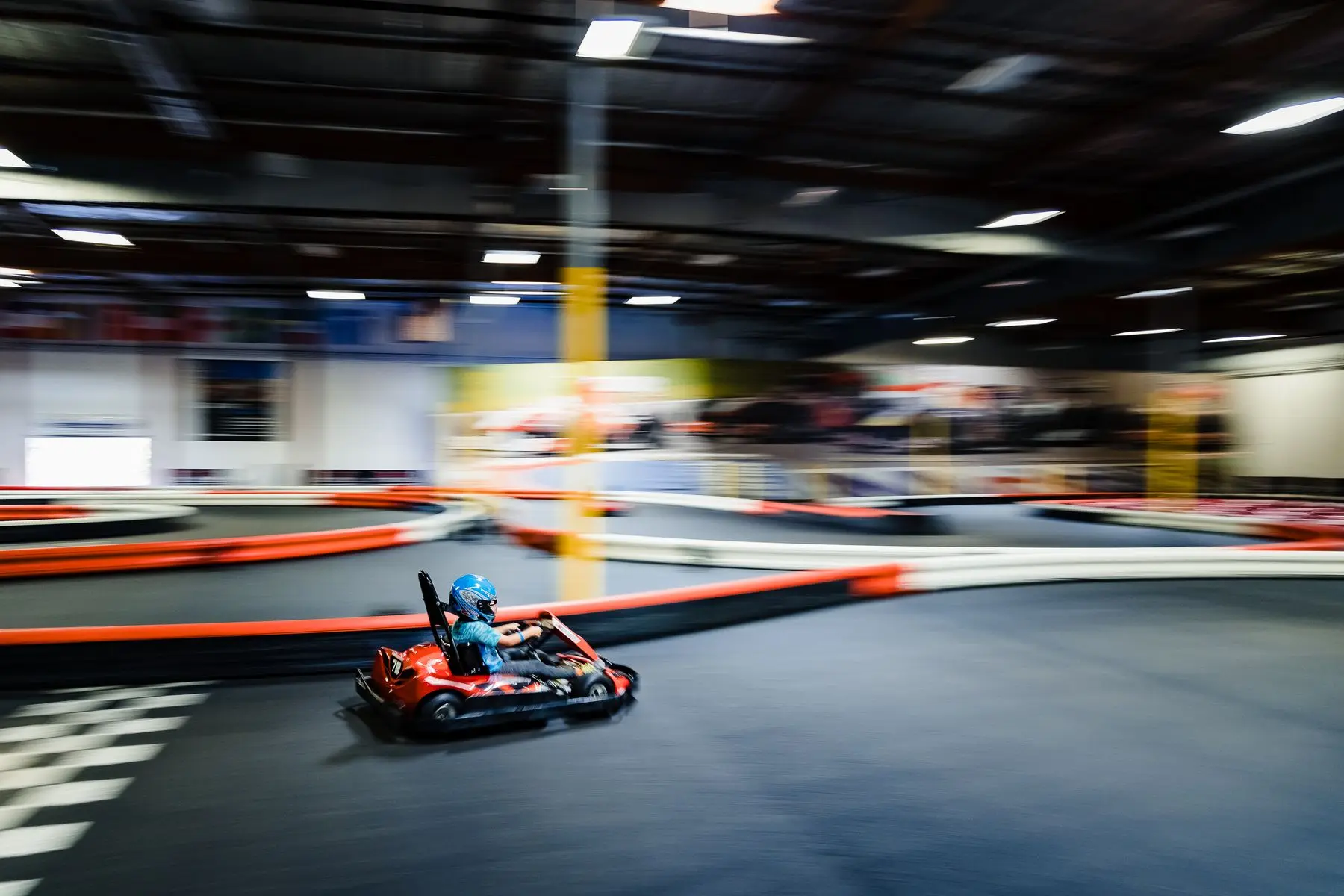 Un enfant casqué file en kart rouge sur une piste couverte aux bordures orange.