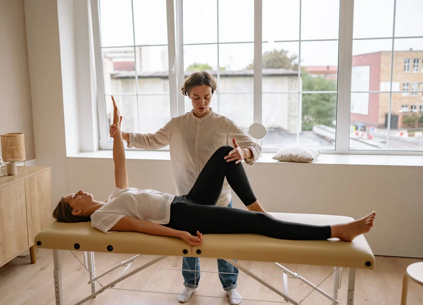 Une kinésithérapeute mobilise le bras et la jambe d'une femme allongée, dans une salle lumineuse.