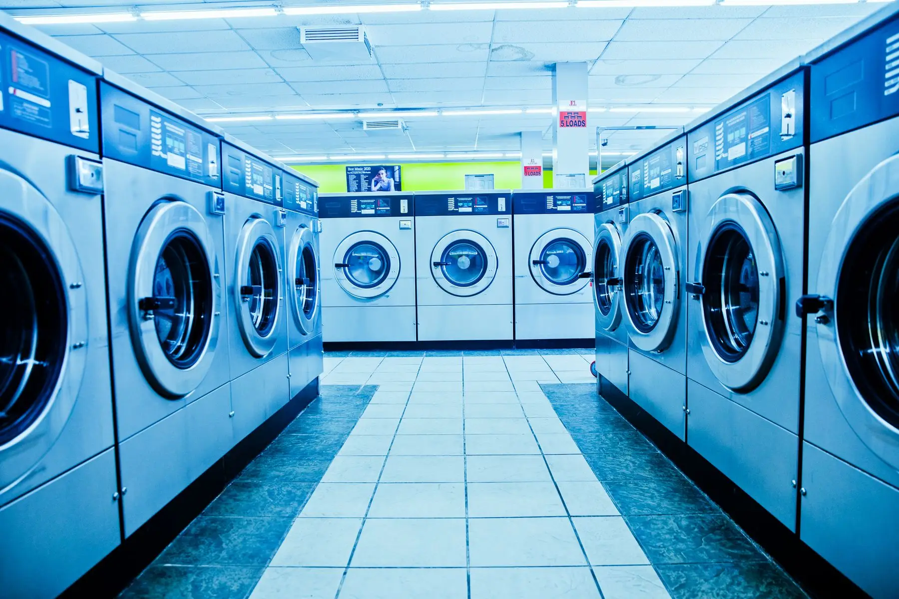 Allée d'une laverie automatique, bordée de rangées de lave-linge à hublot sous un éclairage vif.