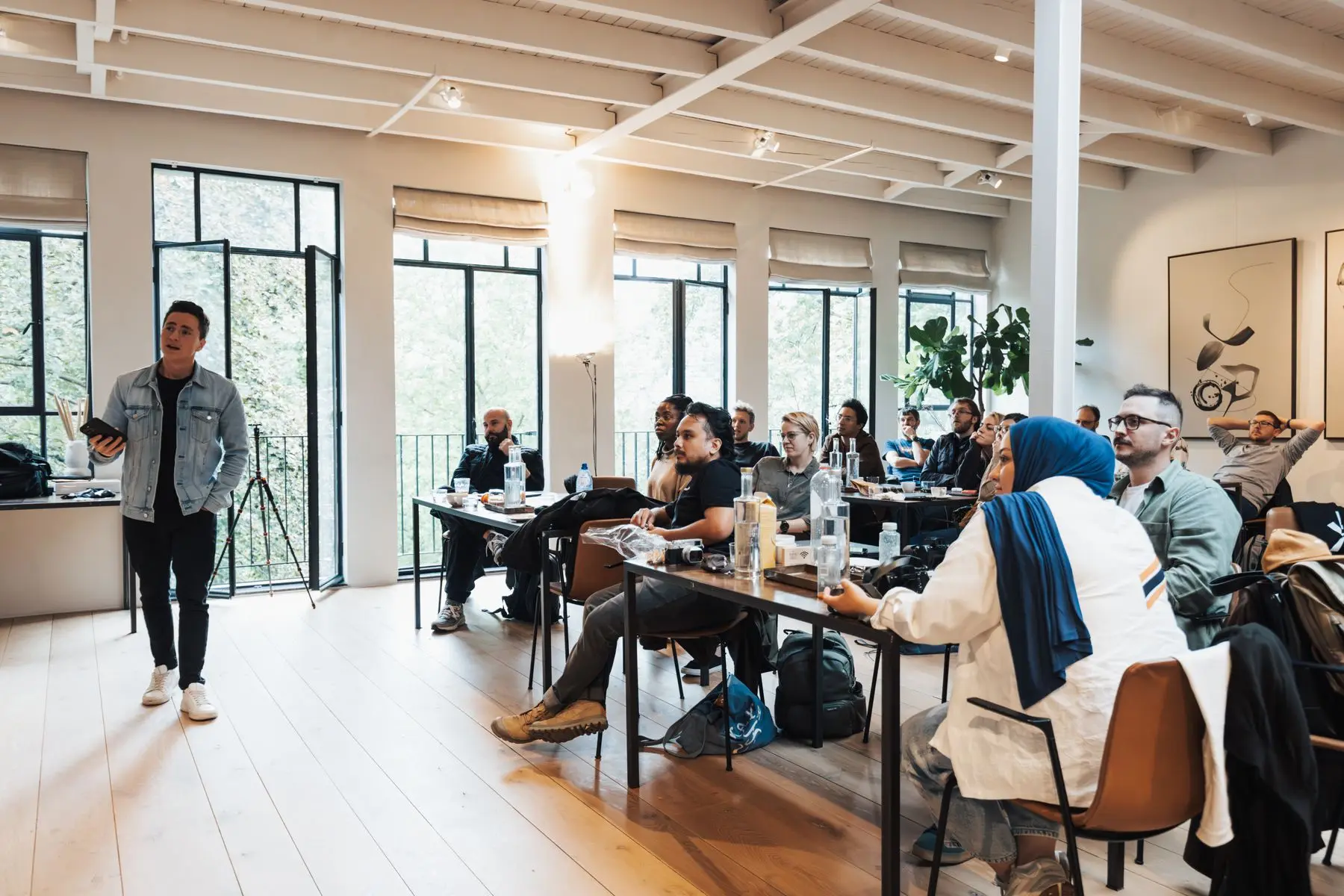 Un formateur debout s'adresse à des adultes assis à des tables, dans une salle lumineuse.