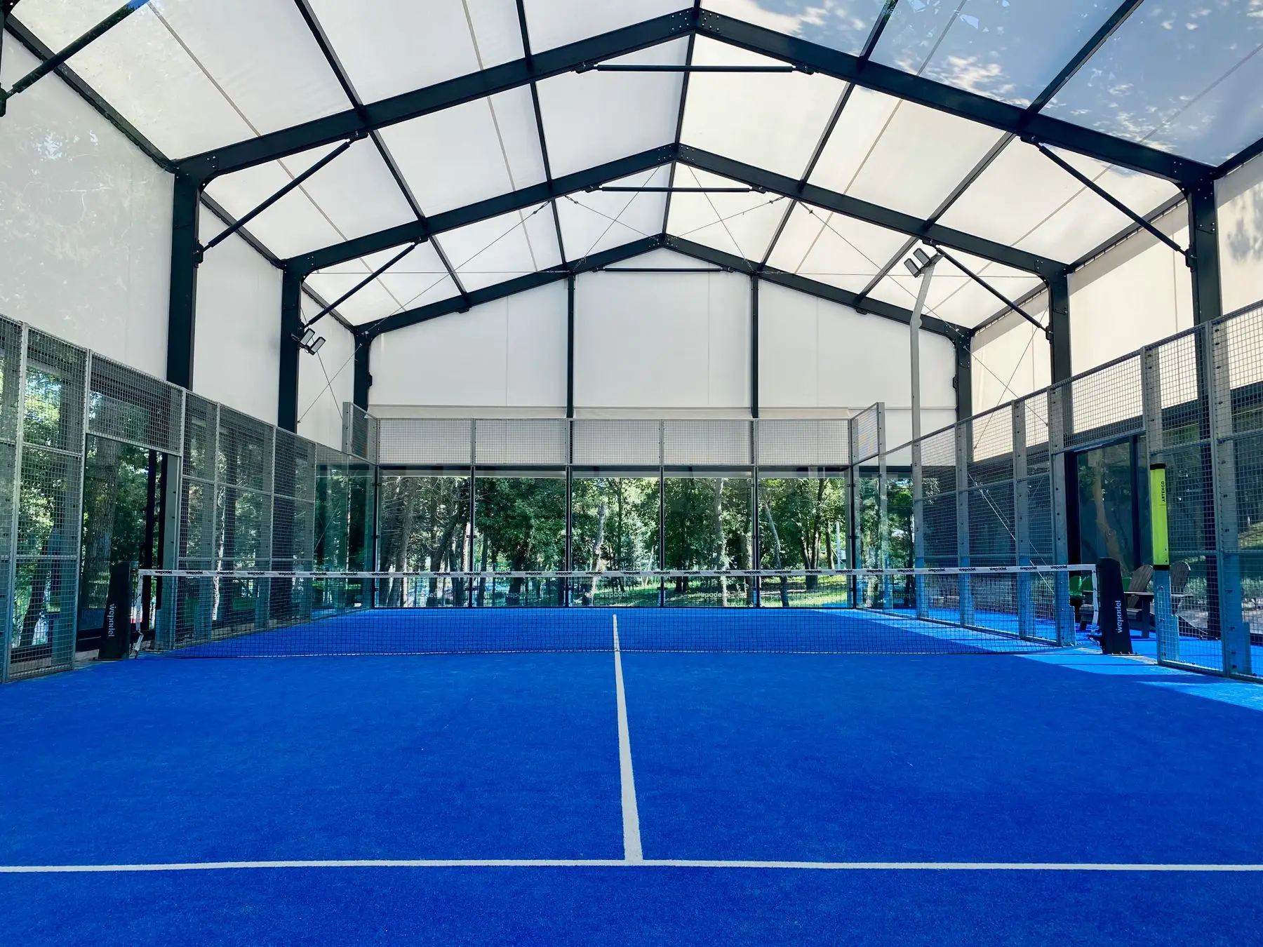 Court de padel au sol bleu, sous une grande halle à toile claire.
