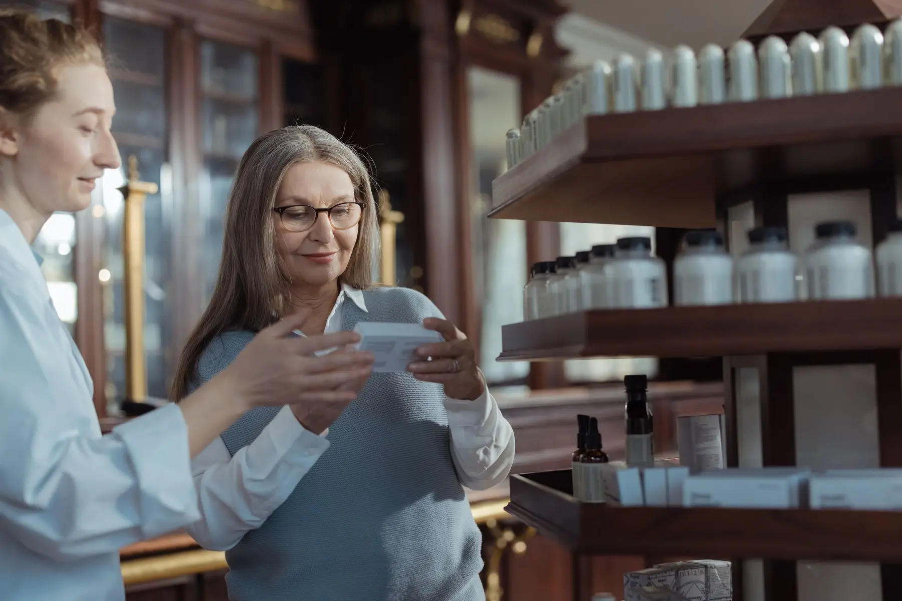 Une pharmacienne conseille une cliente devant des étagères en bois garnies de flacons blancs.